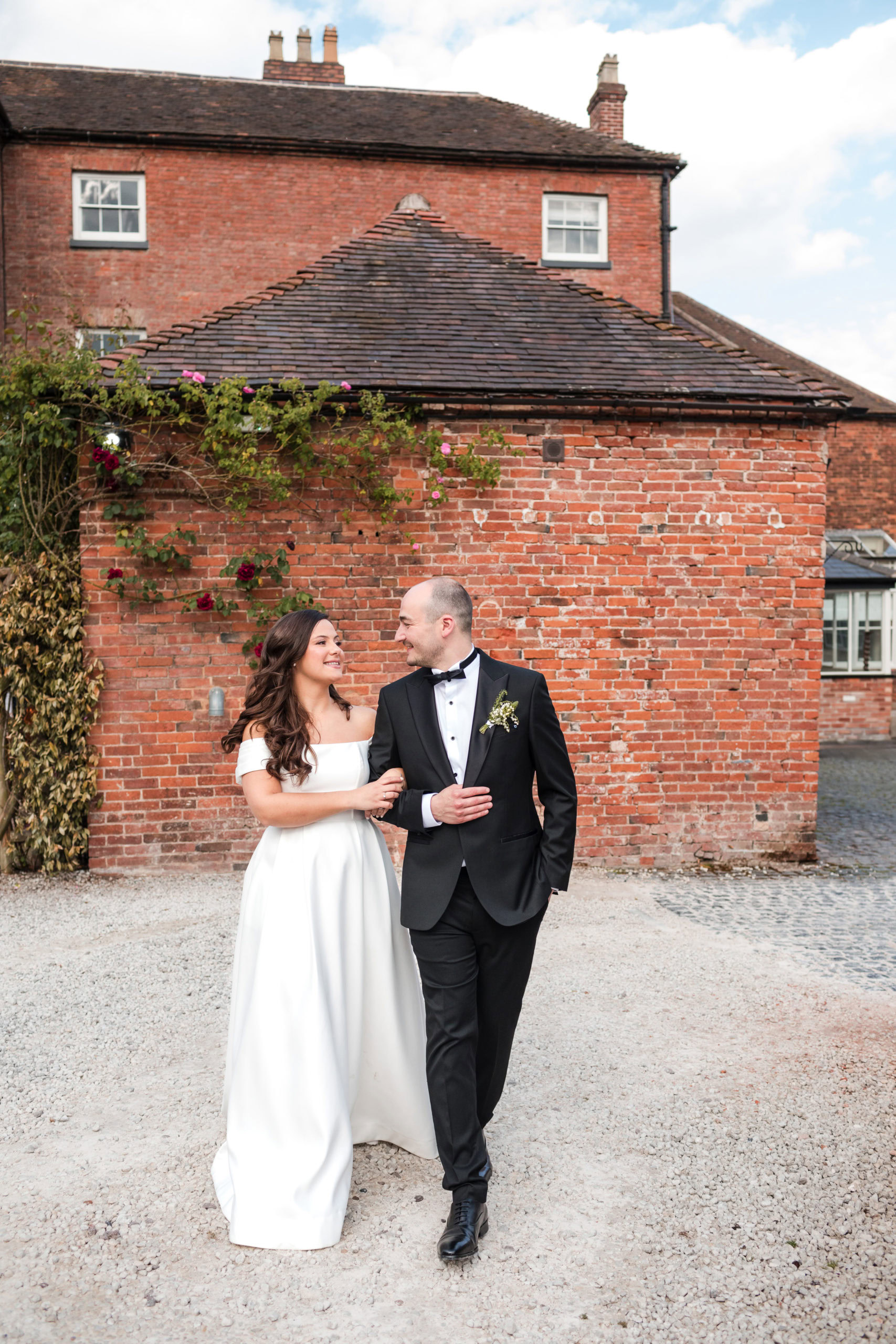 Groom wearing black tie walking with his Bride in front of a rustic brick wall at St Johns House 