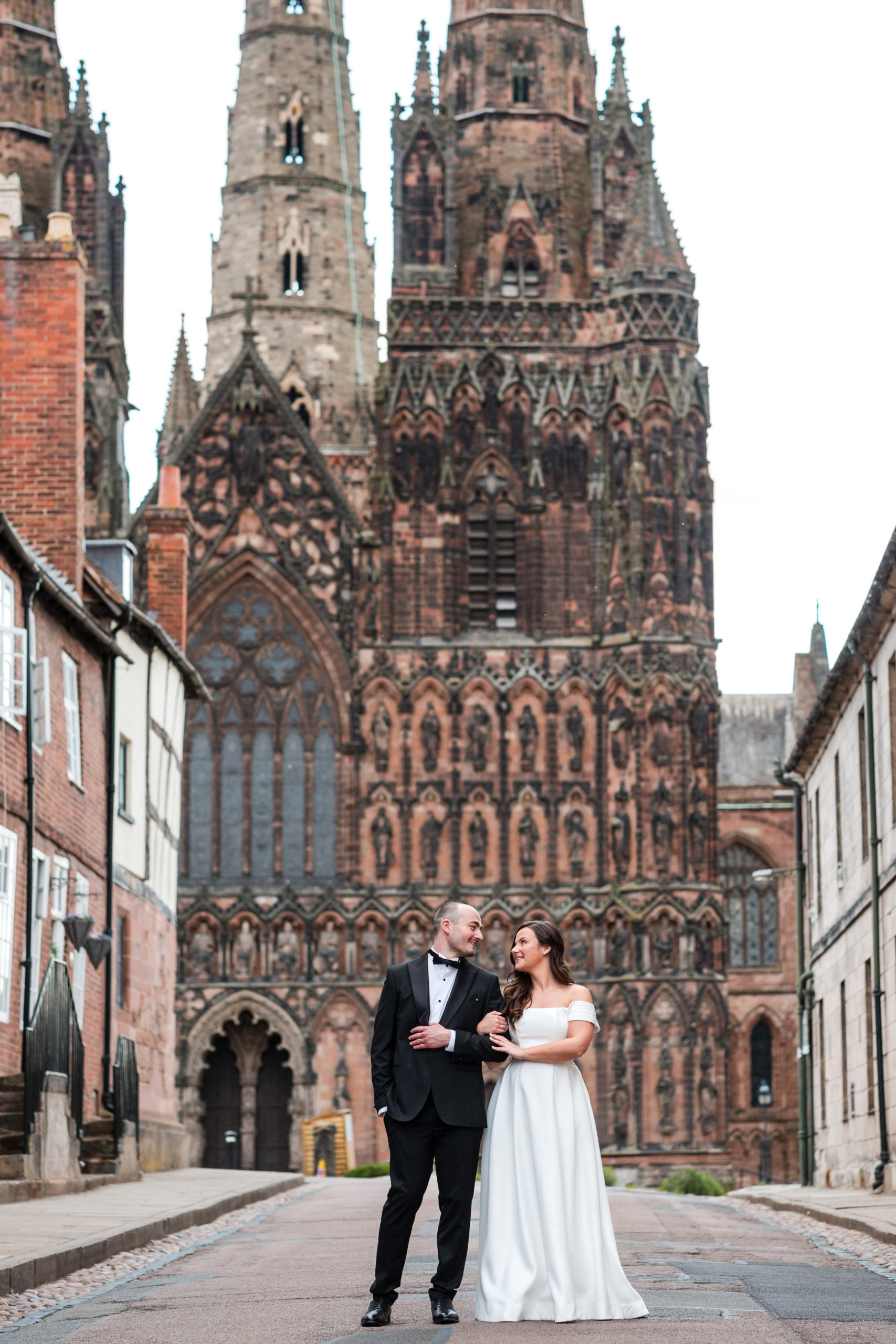 Bride wearing off the shoulder dress links arms with her new husband who wears a tuxedo in front of Lichfield Cathedral after their Lichfield wedding 