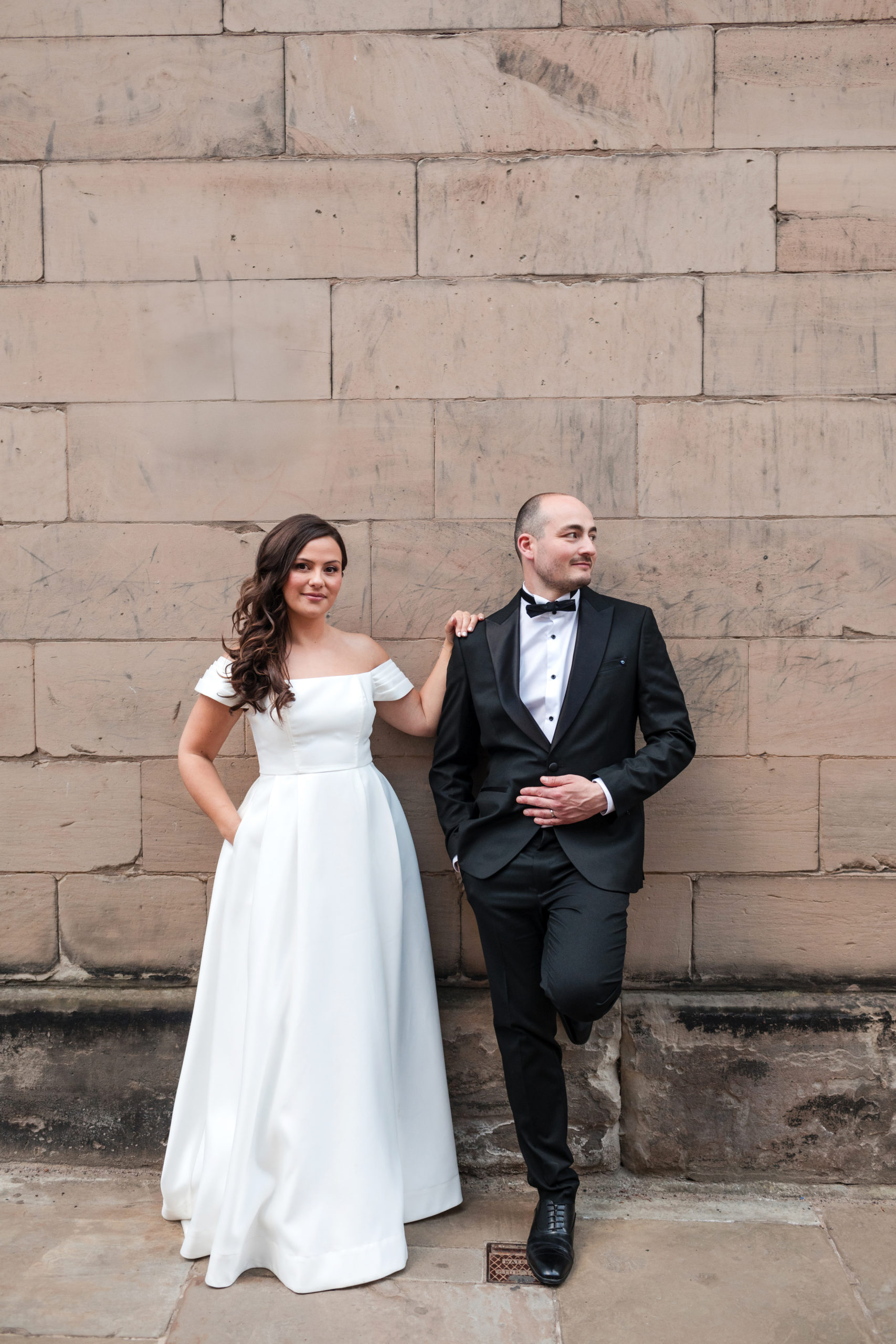 Bride and groom standing against a sandstone wall in Lichfield during their wedding day. The bride wears a classic off-the-shoulder white gown and the groom is dressed in a black tuxedo